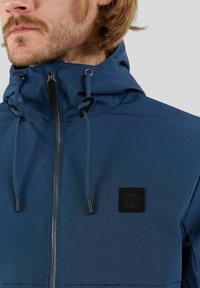 Chaqueta impermeable azul con un cuello alto, capucha ajustable, cierre de cremallera y parche de logo negro en el pecho. Textura suave, diseño minimalista.