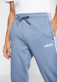 Modré tepláky vyrobené z měkké látky mají stahovací pas, boční kapsy a tři bílé pruhy na bocích s logem Adidas.