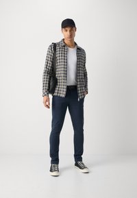Camisa xadrez em verde, azul-marinho e creme, usada sobre uma t-shirt branca; calças azul-marinho e ténis pretos com sola branca; boné preto e mochila.
