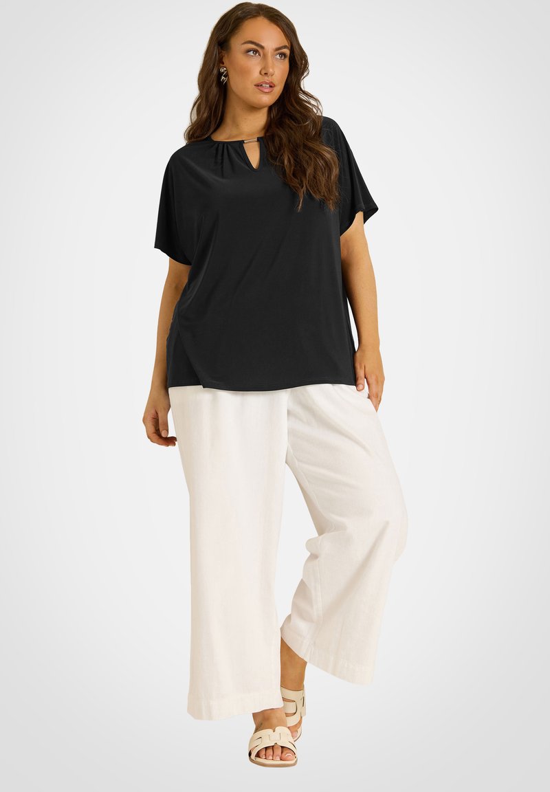 Femme portant un haut noir à manches courtes avec un col rond ajouré, un pantalon blanc à jambes larges et des sandales beiges, debout devant un fond neutre.