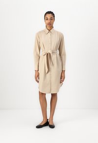 Abito camicia beige con chiusura a bottoni frontale, maniche lunghe e cintura in vita. Tessuto morbido, vestibilità rilassata e spacchi laterali per facilitare i movimenti.