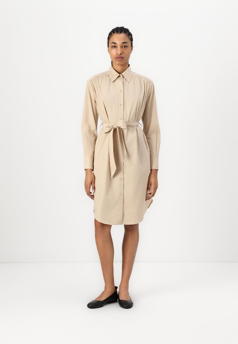 Abito camicia beige con chiusura a bottoni frontale, maniche lunghe e cintura in vita. Tessuto morbido, vestibilità rilassata e spacchi laterali per facilitare i movimenti.