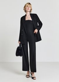 Schwarzer Nadelstreifenanzug mit einem taillierten trägerlosen Oberteil und weit geschnittenen Hosen, kombiniert mit einer schwarzen Handtasche aus Kunstfell und Stöckelschuhen.