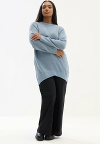 Suéter de punto azul claro con un dobladillo asimétrico y textura acanalada, combinado con pantalones de campana negros y zapatos negros tipo slip-on.