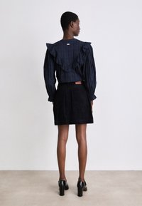 Marineblaue Rüschentbluse mit langen Ärmeln und strukturiertem Design, kombiniert mit einem schwarzen Cordrock. Schwarze Absatzschuhe runden das Outfit ab.