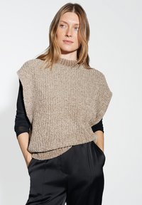 Donna con capelli castano chiaro che indossa un maglione senza maniche beige a trama sopra una maglietta nera a maniche lunghe e pantaloni neri, con le mani in tasca.