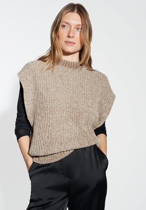 Vrouw met lichtbruin haar die een beige, gewatteerde mouwloze trui over een zwart langhangend shirt en zwarte broek draagt, met de handen in haar zakken.