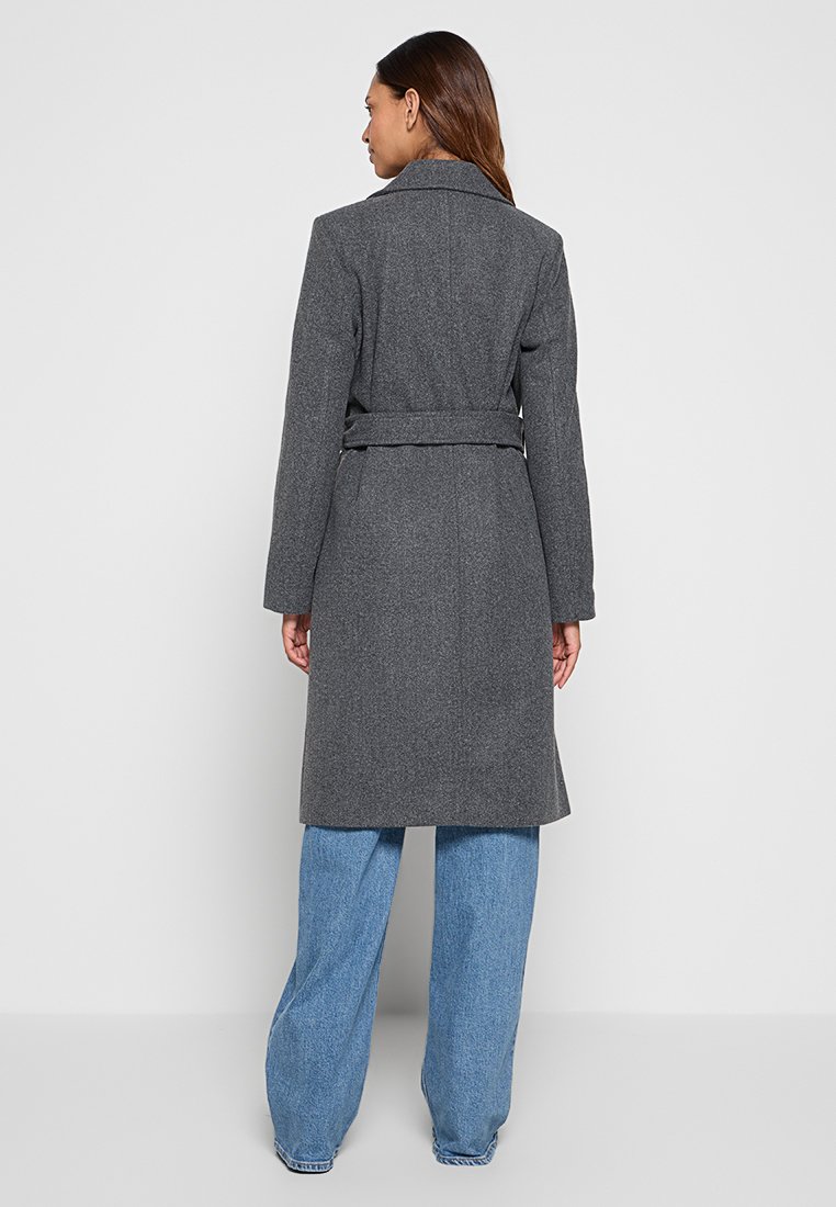 Femme portant un manteau gris ceinturé, longueur genou, et un jean bleu à jambe large, se tenant de dos devant un fond clair.