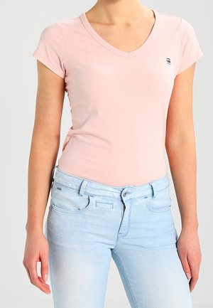 Kobieta ubrana w jasnoróżowy T-shirt z krótkim rękawem i dekoltem w serek oraz jasnoniebieskie, wysokie dżinsy na jednolitym tle.