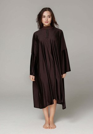 Femme debout pieds nus portant une robe midi ample à manches longues de couleur marron foncé avec une fermeture éclair à l'avant, sur un fond uni.
