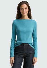 Turkooise longsleeve top met een bootneckline, een gladde textuur en een stiksel dat verticaal over de voorkant loopt, gedragen met donkere jeans.