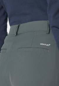 Šviesiai pilkos kelnės su lygiu paviršiumi, turinčios užpakalinius kišenes ir mažą baltą logotipą. Suderintos su tamsiai mėlynu ilgomis rankovėmis viršutiniu drabužiu.