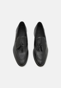 Mocasines de piel negra con detalle de borlas, textura suave, puntera redondeada y acentos de costura. Diseño elegante y suela plana.