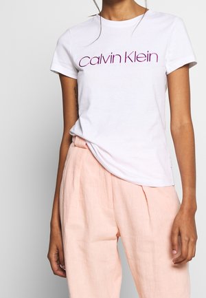 T-shirt blanc en coton avec le texte "Calvin Klein" en violet. Design simple à col rond, manches courtes, tissu léger, coupe légèrement ample.