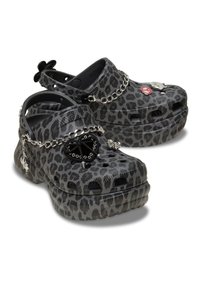 Schwarze und graue Clogs mit Leopardenmuster, Plattformsohle, Belüftungslöcher, Ketten und dekorativen Charms, die eine gemischte Texturierung aufweisen.