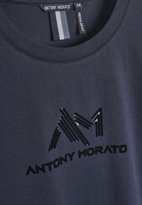 Camiseta azul marino de Antony Morato con el logotipo geométrico negro "AM" y el nombre de la marca debajo, mostrando etiquetas en el cuello etiquetadas como "M" y "Ajuste Regular".