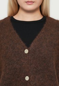Nahaufnahme einer Person, die eine flauschige braune Strickjacke mit zwei hellen Knöpfen über einem schwarzen Rippoberteil trägt, wobei das untere Gesicht und der Hals sichtbar sind.