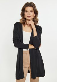 Zwarte lange cardigan van zachte gebreide stof, met een open voorkant, middellange mouwen, gecombineerd met een witte top en beige broek.