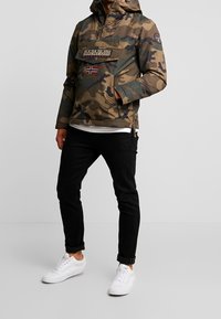 Sudadera con estampado de camuflaje en tonos verdes y marrones, que cuenta con un bolsillo frontal con marca y un detalle de bandera, combinada con jeans negros y zapatillas blancas.