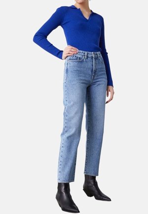 Vrouw draagt een felblauw shirt met lange mouwen, lichtblauwe hoge taille jeans en zwarte enkellaarsjes met spitse neus, staat met één hand op haar heup.