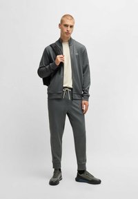 Giacca grigia con zip e colletto alto, abbinata a pantaloni sportivi grigi coordinati, entrambi con una texture liscia e dettagli di design discreti.