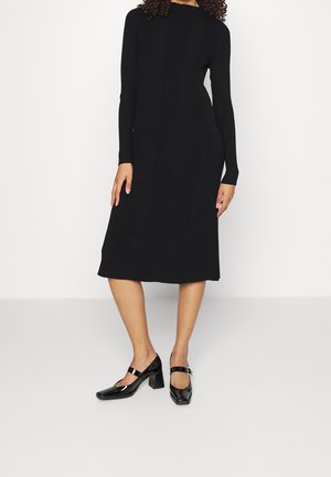 Abito nero lavorato a maglia con maniche lunghe, texture a coste, orlo fino al ginocchio, abbinato a scarpe nere lucide con tacco e cinturino alla caviglia.