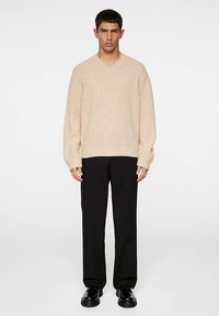 V-nyakú, bézs színű, bordázott mintájú kötött pulóver, egyenes szabású fekete nadrággal és fekete cipővel, egy férfi modell által viselve, egyszínű háttér előtt.