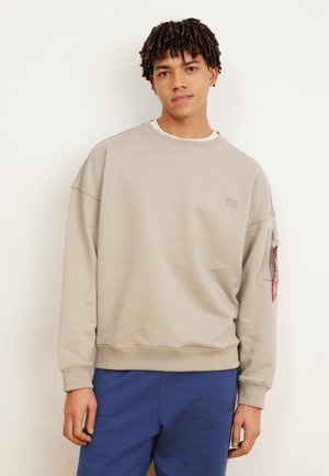 Junger Mann mit kurzen Dreadlocks, der einen beigen Pullover mit Reißverschlusstasche und rotem Etikett am Ärmel sowie blaue Hosen vor einem einfarbigen Hintergrund trägt.