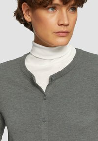 Knopen cardigan van grijs breiwerk, met een V-hals en twee donkere knopen. Gelaagd over een witte turtleneck, zachte textuur zichtbaar.