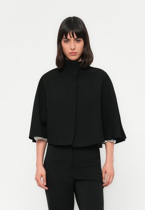 Woman with straight dark hair wearing a black high-neck cape jacket and matching black pants against a white background.