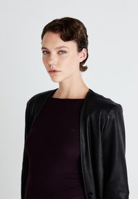 Giacca di pelle nera con texture morbida e dettagli con bottoni, indossata sopra un top fitted a coste di colore viola scuro. Presenta un collo rotondo e un logo.