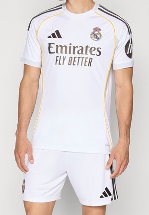 Mand iført hvid Real Madrid fodboldtrøje med gule piping, sorte Adidas-striber og Emirates Fly Better sponsorlogo.