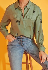 Green, long-sleeve shirt with two chest pockets and embroidered plant patterns. Paired with light blue distressed jeans, sitting on a stool.