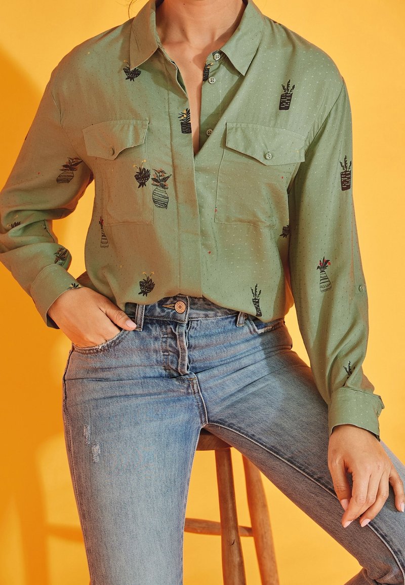 Green, long-sleeve shirt with two chest pockets and embroidered plant patterns. Paired with light blue distressed jeans, sitting on a stool.
