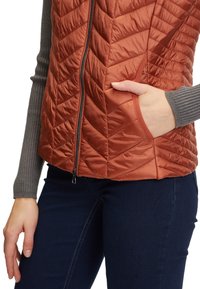 Zbliżenie na osobę w rdzawym, pikowanym kamizelce z zamkiem błyskawicznym z przodu, w niebieskich dżinsach i szarej, prążkowanej koszuli z długim rękawem, z prawą ręką w kieszeni kamizelki.