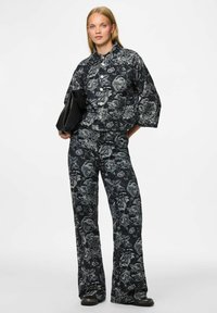 Vrouw in bijpassende zwarte en grijze bloemen wijdere broek en knopenjack, met een zwarte gestructureerde clutch in haar hand, staande tegen een witte achtergrond.