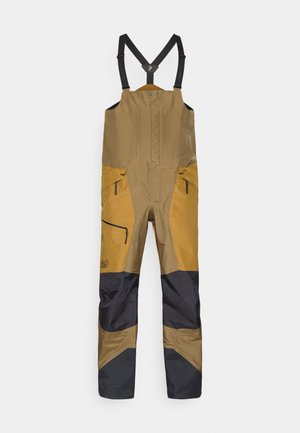 Brune og sorte bib-overaller med justerbare sorte stropper og lynlåslommer, designet til udendørs eller arbejdsmæssig brug.