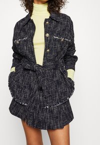 Veste en tweed marine avec des boutons dorés et une taille ceinturée, associée à un col roulé vert clair. Tissu texturé avec des bords effilochés.