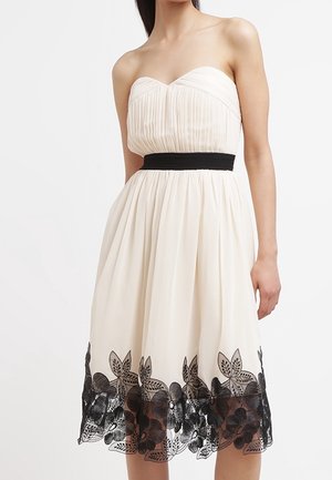 Strapless cream dress with pleated bodice and a black waistband. Skirt features intricate black floral lace trim at the hem.