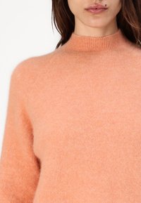 Pulover moale, cu guler rotund, într-o nuanță deschisă de piersică, realizat dintr-un material pufoș, cu o textură delicată și guler cu nervuri. Croială casual.