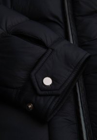 Gros plan sur une doudoune noire présentant une texture matelassée, un bouton pression argenté et une fermeture éclair le long du bord. Matière douce et lisse.