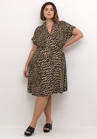 Robe à imprimé léopard marron et noir, manches courtes, col en V, taille cintrée, jupe fluide. Associée à des talons épais noirs.