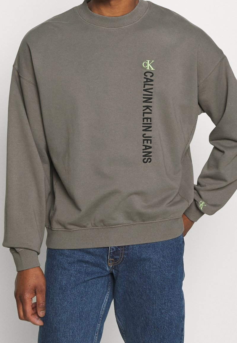 Sudadera gris de Calvin Klein con mangas largas, puños y dobladillo de canalé. Presenta texto vertical en negro y un pequeño logo verde en el pecho.