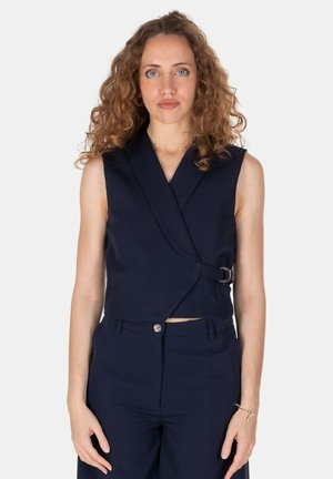 Donna con capelli ricci che indossa un gilet avvolgente smanicato blu navy e pantaloni a vita alta coordinati su uno sfondo bianco semplice.
