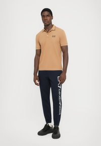 Beige polo skjorte med svarte kragedetaljer og logo, kombinert med marineblå joggebukser med hvit branding, og svarte joggesko.