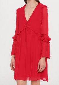 Rode chiffon jurk met een diepe V-hals, gerimpelde afwerking en een gather taille. Lange mouwen hebben elastische boorden en een ontspannen, vloeiende silhouet.