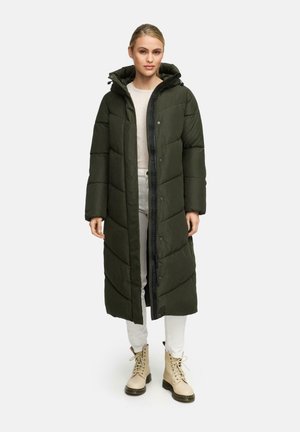 Lange, met capuchon gevoerde donzen jas in donkergroen, met een doorgestikt patroon en drukknoopsluiting, gecombineerd met lichtgekleurde broek en beige enkellaarsjes.