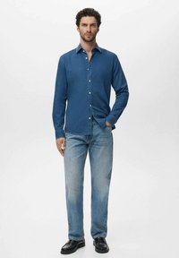 Mann steht da, trägt ein blaues Hemd mit Knöpfen, hellblaue Jeans und schwarze Schuhe, mit einer Hand in der Tasche.