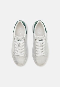 Sølv sneakers med en teksturert overflate, hvite snører og grønne detaljer på hælen. Merking og størrelse er synlige inni skoen.