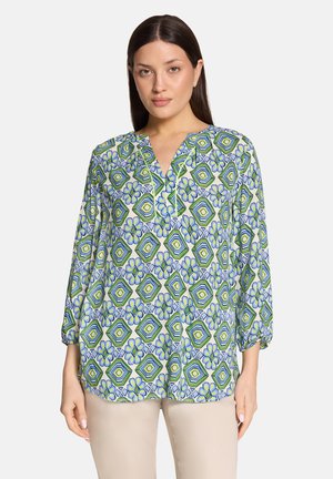 Frau trägt eine langärmelige Bluse mit grünen, blauen und weißen geometrischen sowie floralen Mustern, kombiniert mit beigen Hosen.